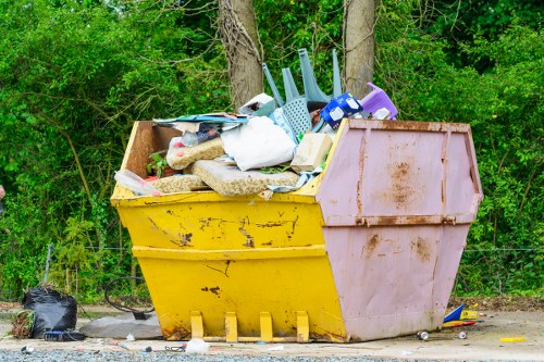 A large two-tone skip, with a yellow base and pink upper section, is positioned on a gravel surface beside a backdrop of green trees and bushes. The skip is overflowing with various household waste items, including a white plastic bag, a vacuum cleaner, plastic buckets, and other miscellaneous rubbish. Some objects, such as a purple container and a blue plastic scoop, are stacked or leaning against the debris. The skip shows signs of dirt and weathering, with visible grime and rust marks on its surfaces. To the left of the skip, a small black plastic bag lies on the ground. The surrounding environment appears to be an outdoor area, possibly a driveway or service access point, with natural greenery behind it. Office Clearance Plumstead offers rubbish removal services suitable for clearing waste like this in the local area, including Plumstead and nearby districts, supporting efficient and responsible waste disposal.