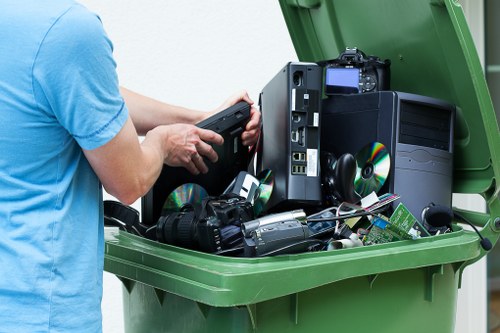 A person wearing a light blue T-shirt is seen from the side, using a black handheld device to manually compress electronic waste inside a large green plastic skip. The skip contains various discarded electronic items, including a black desktop computer tower positioned upright with a visible DVD drive and power supply, several black computer mice, and a digital camera with a black body and attached strap. Some CDs or DVDs are scattered around, and the background shows an outdoor setting with pavement or driveway, suggesting a rubbish removal or clearance operation in progress. This scene exemplifies the disposal of electronic waste as part of a professional rubbish clearance service, such as that offered by Office Clearance Plumstead, which operates in the Plumstead area.