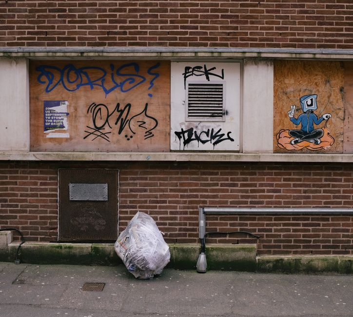 A tall, rectangular yellow needle drop box positioned against a dark, textured wall in an urban environment. The box is made of metal with a slightly weathered finish, including visible rust and dirt 