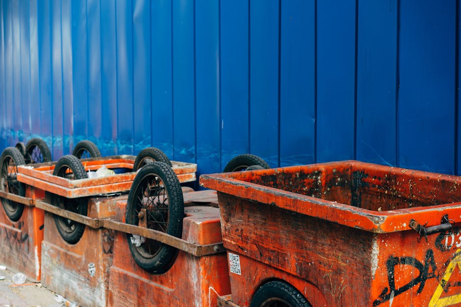 A row of large orange plastic skip bins with black rubber wheels situated along a driveway or paved surface, positioned next to a tall blue metal fence. The bins appear weathered, with visible dirt, g