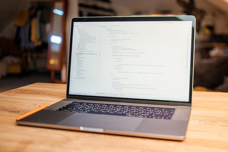 A close-up view of a laptop computer screen displaying lines of JavaScript code with syntax highlighting, variables, and functions, situated on a desk or workspace. In the background, a blurred citysc